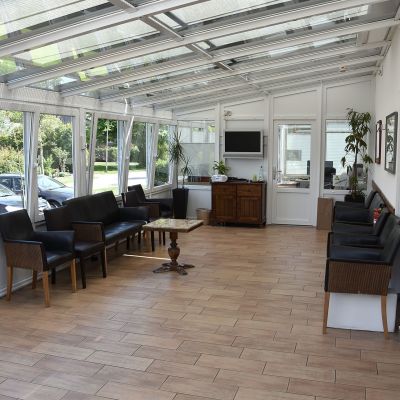 Ein heller, langer Warteraum mit Glasdach und vielen Fenstern. Dunkle Stühle und Tische sind an den Wänden aufgestellt. Ein Fernseher und einige Pflanzen sind ebenfalls zu sehen. Der Boden besteht aus Holzfliesen.