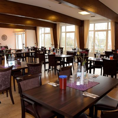Ein Speisesaal mit dunklen Holztischen und -stühlen, die in Reihen angeordnet sind. Auf den Tischen stehen Flaschen und Gläser. Große Fenster lassen viel Tageslicht herein. Die Einrichtung wirkt ordentlich und einladend.
