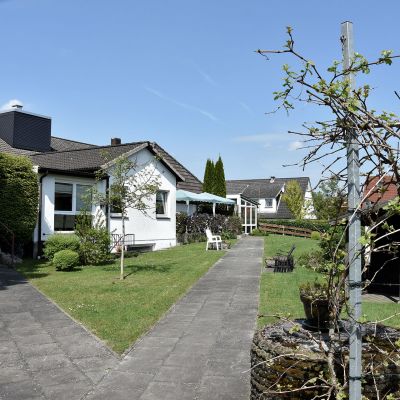 Das Bild zeigt einen gepflegten Gartenbereich mit einem gepflasterten Weg, der zu einem Wohnhaus führt. Links und rechts des Weges sind Rasenflächen und Sträucher. Im Hintergrund stehen Gartenmöbel auf einer Terrasse. Der Himmel ist blau.