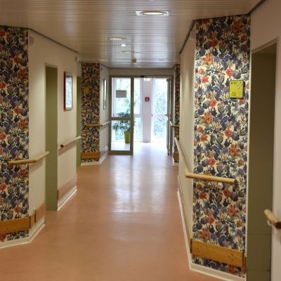 Ein Flur mit rosa Boden, weißen Wänden und bunten blumengemusterten Tapeten an Abschnitten. An den Wänden sind Holzhandläufe befestigt. Am Ende des Flurs befindet sich eine Glastür mit Blick nach draußen.