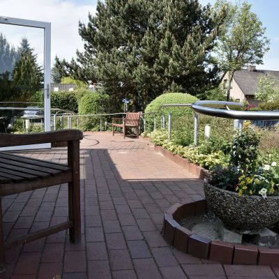 Eine Terrasse mit rotem Ziegelboden und Metallgeländer führt zu einem Holzstuhl. Links steht ein Holztisch mit Bank. Rechts sind Blumen in einem Pflanzgefäß, dahinter Bäume und ein Haus. Der Himmel ist blau.