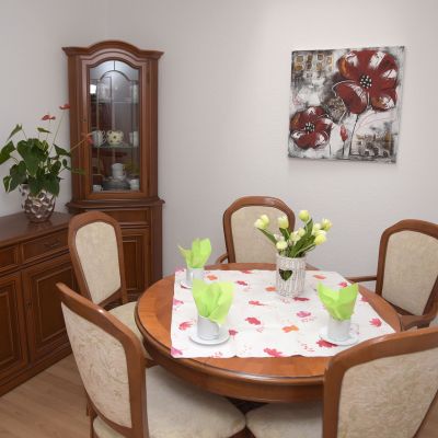 Ein Esszimmer mit einem runden Holztisch und vier Stühlen. Auf dem Tisch stehen Tassen, grüne Servietten und weiße Blumen. Links steht ein Schrank mit Geschirr und Pflanzen, an der Wand hängt ein Bild mit roten Blumen.