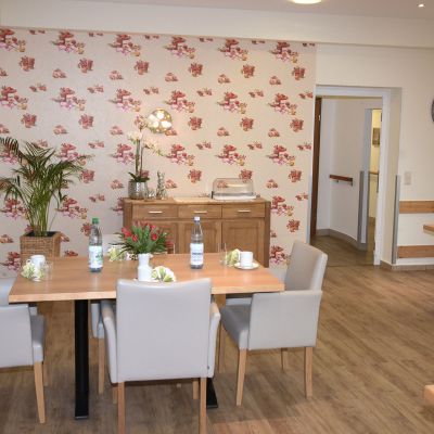 In einem gemütlichen Raum stehen zwei Tische mit beige gepolsterten Stühlen. Auf den Tischen sind Blumen und Wasserflaschen. Eine Kommode mit Lampe und Uhr an der Wand vervollständigen das Bild. Der Boden ist aus Holz.