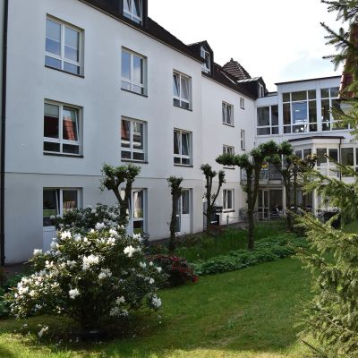 Ein dreistöckiges weißes Wohngebäude mit Dachgauben und großen, rechteckigen Fenstern. Der gepflegte Garten im Vordergrund hat geschnittene Bäume, Büsche und einen grünenden Rasen. Ein Wintergarten ist an der rechten Seite angebaut.