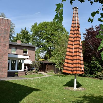 Ein Garten mit einem geschlossenen, orange-gestreiften Sonnenschirm in der Mitte. Im Hintergrund ein Backsteinhaus mit Terrasse. Umgeben von Bäumen und Sträuchern. Der Himmel ist klar und blau.