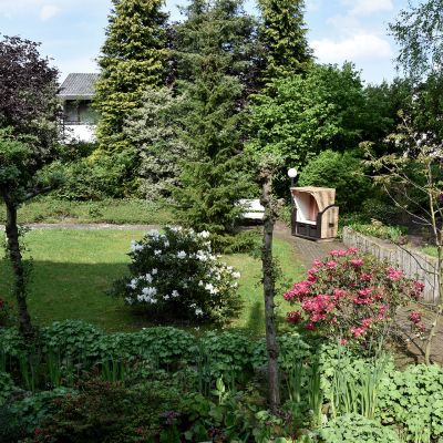 Ein Garten mit grüner Rasenfläche und bunten Blumenbeeten. Einige Bäume und Büsche umgeben die Wiese. Im Hintergrund steht ein weißes Haus, verdeckt von hohen Bäumen. Ein Strandkorb steht auf einem gepflasterten Weg rechts im Bild.