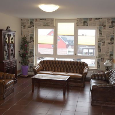 Ein Wohnzimmer mit braunen Ledermöbeln: einem Sofa, zwei Sesseln und einem Couchtisch. Im Hintergrund große Fenster mit halb heruntergelassenen Jalousien, eine Zimmerecke mit einer Pflanze und ein Holzschrank. Wände mit Zeitungsmuster.