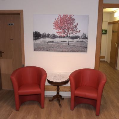 Zwei rote Stühle stehen auf einem Holzboden vor einer weißen Wand. Zwischen den Stühlen befindet sich ein kleiner runder Tisch. Über ihnen hängt ein Bild mit einem Baum und Landschaftsmotiv. Türen führen in angrenzende Räume.