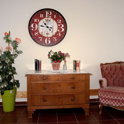 Ein Zimmer mit einer Kommode, auf der Pflanzen und Bücher stehen. Links ist eine große grüne Vase mit Rosen. Darüber hängt eine große Wanduhr. Rechts steht ein gepolsterter, roter Sessel. Der Boden ist gefliest.