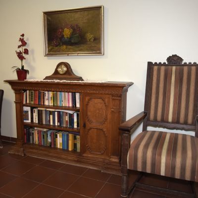 Ein Raum mit braunen Fliesenboden. Links steht ein dunkelsessel und daneben ein Holzbücherregal mit darauf stehender Uhr und Topfpflanze. Rechts ist ein großer Stuhl mit Stoffbezug. An der Wand hängt ein Gemälde.